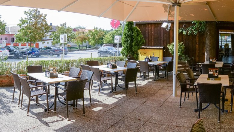 Guest garden, © Siebenbrunnerhof A sunny guest garden with empty tables and chairs, surrounded by plants and a large parasol.
