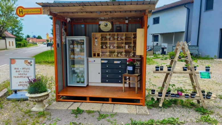 The honey stop, © Imkerei Pell A small roadside stall selling organic products such as honey, eggs and soaps. Next to it are plants on a wooden frame.