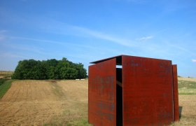 Wind cube house, © StadtGemeinde Mistelbach Rusty metal cube on a field with trees in the background.
