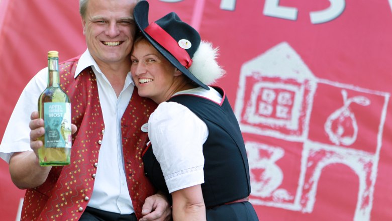 Zarl family, © weinfranz.at A man and woman in traditional dress smile and hold a bottle of apple and pear cider against a red background.