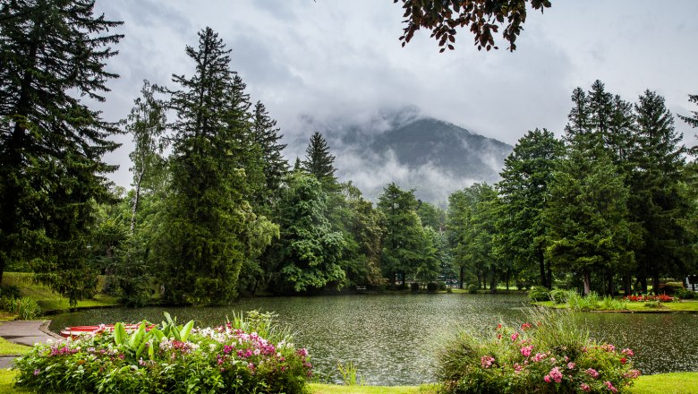 Reichenau Spa Park, © Wiener Alpen/Nadja Meister A picturesque park with a pond, surrounded by trees and flowers, with a misty mountain in the background.