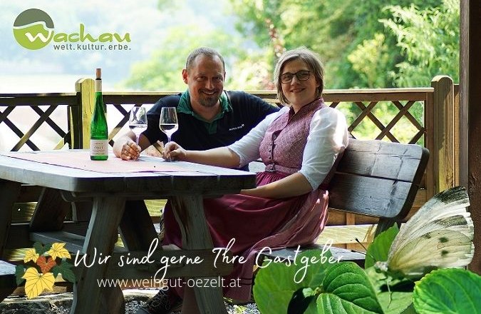 Özelt-Reischer family, © DM A couple sits at a wooden table outdoors with wine and glasses. Nature can be seen in the background. Text: "We are happy to be your hosts".
