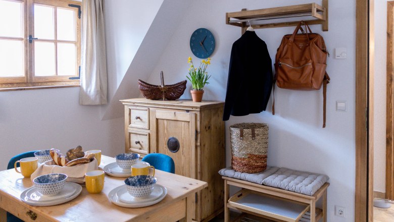 Breakfast corner, © Monika Pölzer Cozy breakfast corner with wooden table, dishes, yellow cups, chest of drawers, jacket, rucksack and flowers on a cupboard.