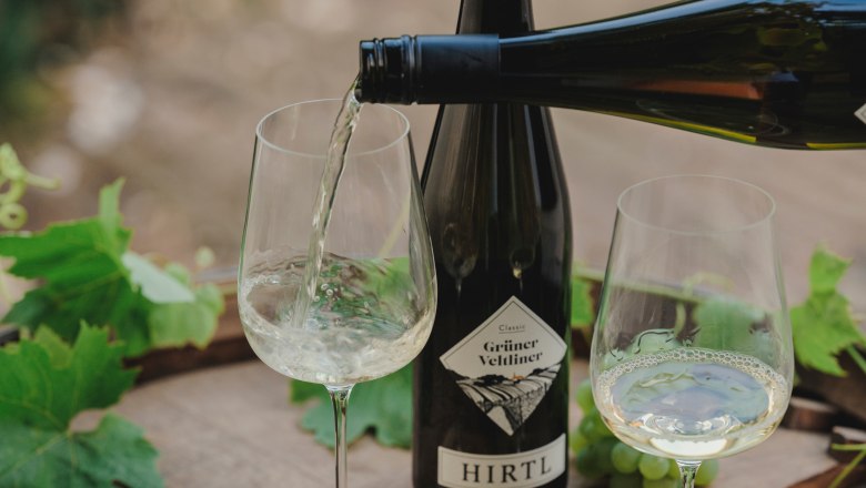 Enjoy wines, © Michael Reidinger White wine is poured into a glass, next to it a bottle of Grüne Veltliner from Hirtl and green grapes on a wooden table.