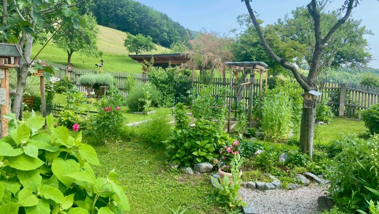 Apartments Riedl, © Wiener Alpen A well-kept garden with flowers, trees and a gravel path, surrounded by a wooden fence and a green meadow in the background.