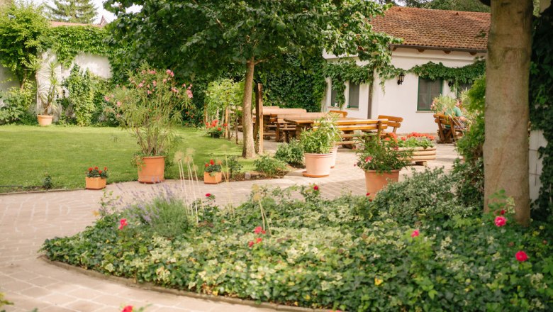 Garden, © Felix Sommer A well-tended garden with lawn, flower beds and terracotta pots. In the background, a house with a terrace and wooden tables.