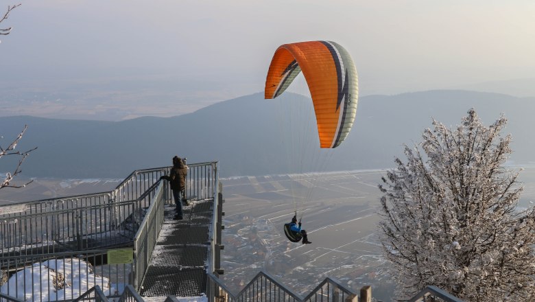 Hohe Wand Nature Park, © Naturpark Hohe Wand/Fraller A paraglider flies over a snow-covered landscape in the Hohe Wand Nature Park, while a person stands on a viewing platform.