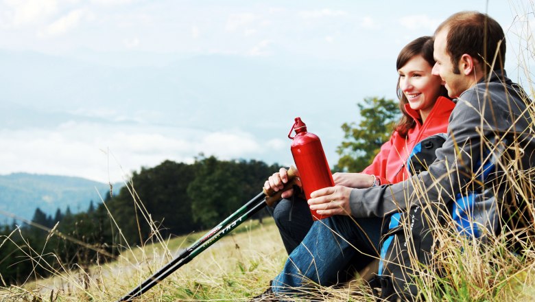 Couple on the Hinteralm, © weinfranz.at A couple sits on a meadow with hiking poles and a red water bottle, mountains and clouds in the background.
