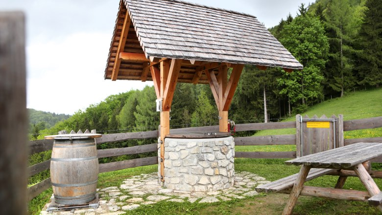 Reinsberg cider fountain, © weinfranz.at A traditional well with a wooden roof and stone wall, next to a wooden barrel and a picnic bench, surrounded by green countryside.