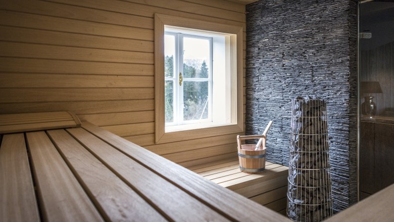 Villa Antoinette, © Matthias Kronfuss Interior view of a sauna with wood paneling, a window, a bucket and a stove with stones.