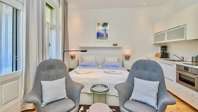 Top 4, © VP FeWo OG Modern studio apartment with double bed, two gray armchairs, kitchenette and large windows.