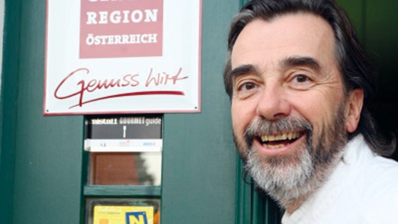 Manfred Buchinger, © Gasthaus Zur Alten Schule A man with a beard smiles in front of a sign reading 'Genuss Region Österreich'.