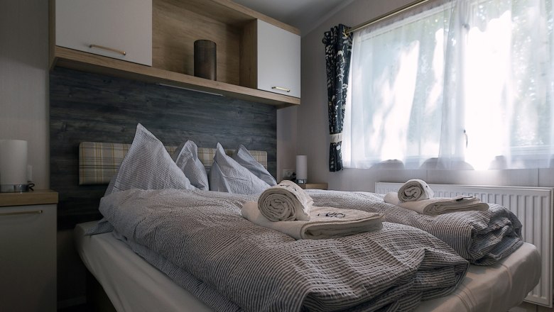 Bedroom, © Leopold Pluschkowitz A cozy bedroom with a double bed, grey bed linen and rolled towels. A window with curtains lets in natural light.