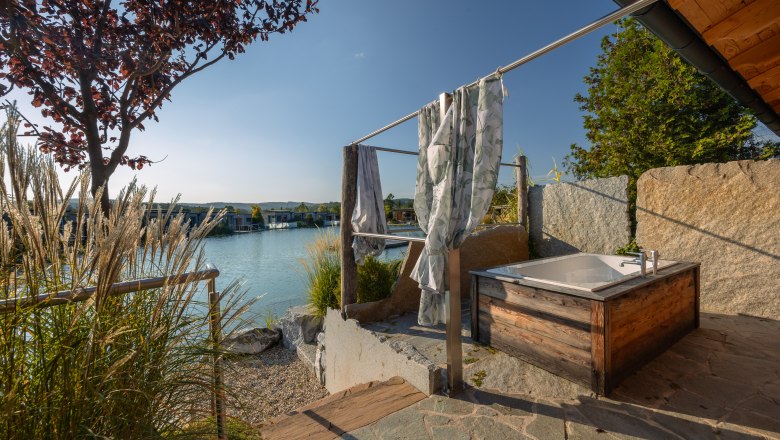 Kittenberger Chalets at the Gartensee, © Niederösterreich Werbung / Maximilian Pawlikowsky Outdoor area with bathtub, view of a lake and chalets in the background.