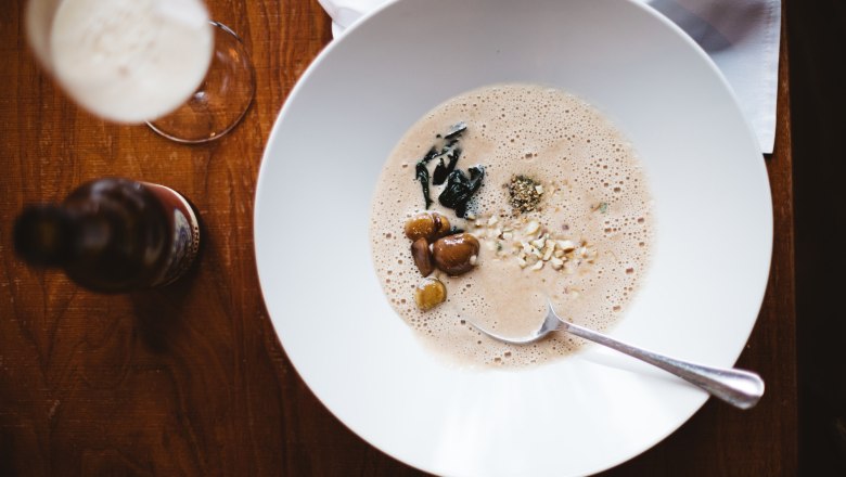 Countless chestnut creations: here chestnut soup, © Niederösterreich-Werbung/Mara Hohla A bowl of chestnut soup with nuts and herbs on a wooden table, a glass and a bottle next to it.