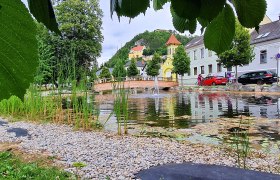 View of the mountain church in Pitten, © Wiener Alpen, Schuh A pond with pebbles and reeds, a bridge and colorful buildings in the background.