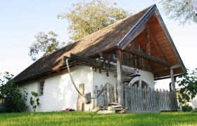 Museum outside, © Fam. Buchinger Traditional building with wooden roof and fence, surrounded by green meadow and trees.