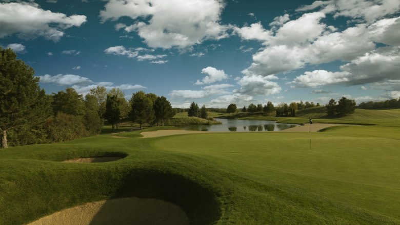 Hole 4, © FONTANA Golf course with sand bunker, green and pond under a blue sky with clouds.