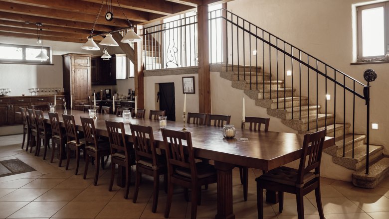 Kitchen and dining table, © Weinakademie Retz Large dining table with many chairs in a rustic room with wooden ceiling and staircase.