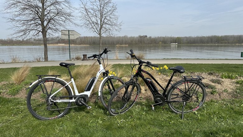 E-bike rental Tulln tourist information, © Stadtgemeinde Tulln Two e-bikes are parked on a meadow in front of a river.
