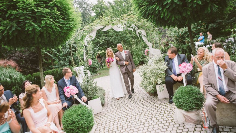 Wedding in the love garden, DIE GARTEN TULLN, © Klaus Bauer Bride and attendant walk through a garden arch, surrounded by guests.