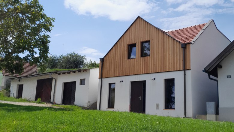Distillery - Kollmann, Destithek, © Brennerei - Kollmann A modern building with a wooden façade and red roof tiles stands on a green hill under a blue sky.