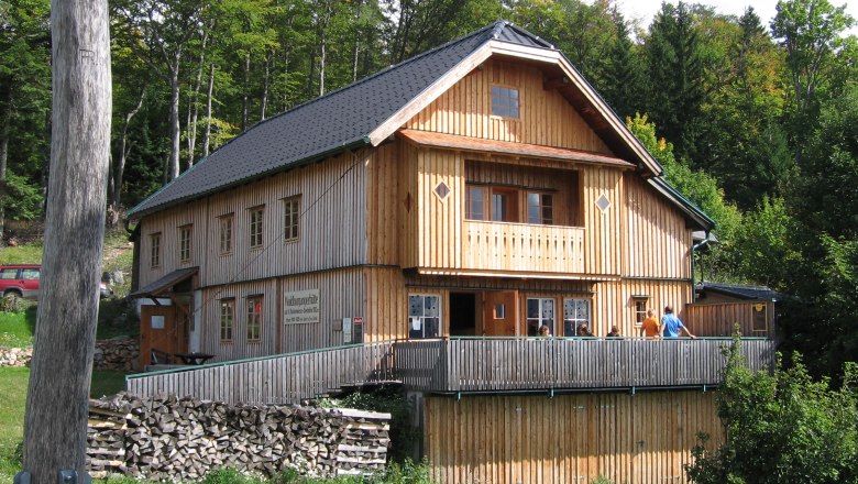 Waldburgangerhütte, © Maria Stroebl A wooden hut in the forest with a terrace and people on it.