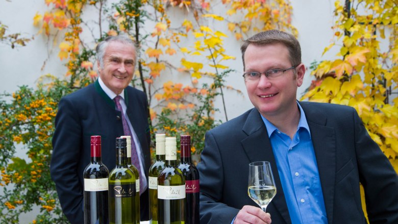 Josef and Christian Pleil, © Fotostudio Semrad Two men stand at an outdoor table with bottles of wine, surrounded by autumnal plants.