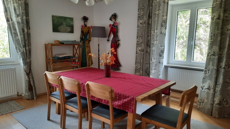 Dining area, © Dr. Eva Dirnbacher Dining area with wooden table, red table runner, wooden chairs, curtains and decorations on the wall.