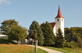Ofenbach parish church, © Thermengemeinden Ofenbach parish church with red tower roof, surrounded by trees and meadows.