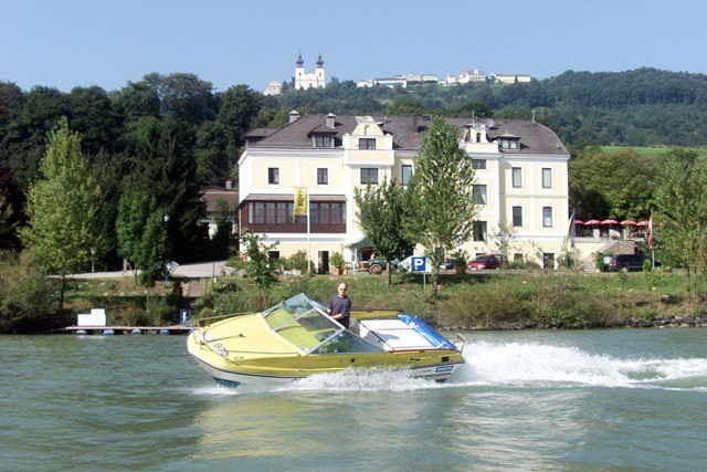 Housewithboat, © Wachauerhof Renner GMBH A yellow motorboat sails on a river in front of a large, light-colored building with hills in the background.