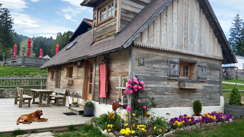 Panoramic hut on the Mönichkirchner Schwaig, © Wiener Alpen A rustic wooden hut with a flower bed and a dog on the terrace, surrounded by green countryside and red parasols.