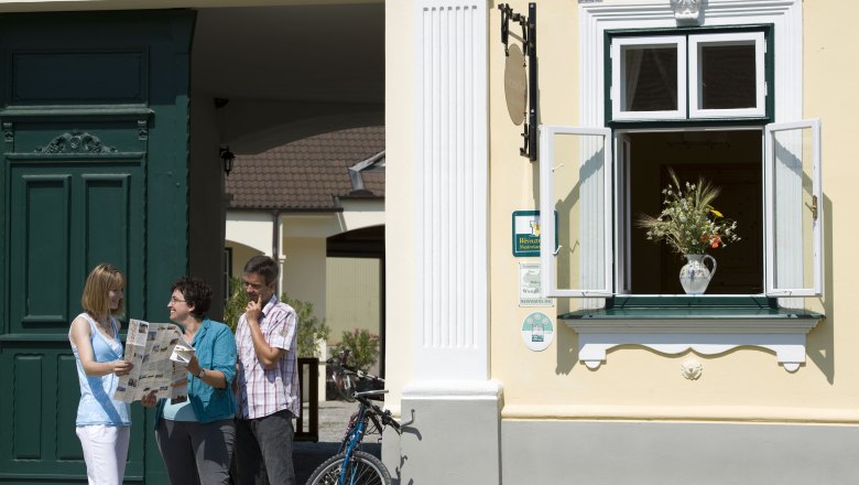 Winzerhof & Guesthouse Greil, © M. Greil Three people are standing in front of a yellow building with a green door, reading a card. A window with a bouquet of flowers is open.