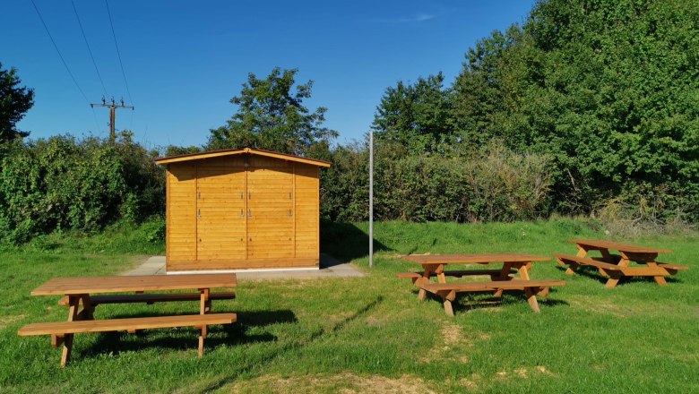 Bachleiten Platzl, © Winzerhof Ernst Wooden hut with picnic tables on a meadow, surrounded by trees and bushes under a blue sky.