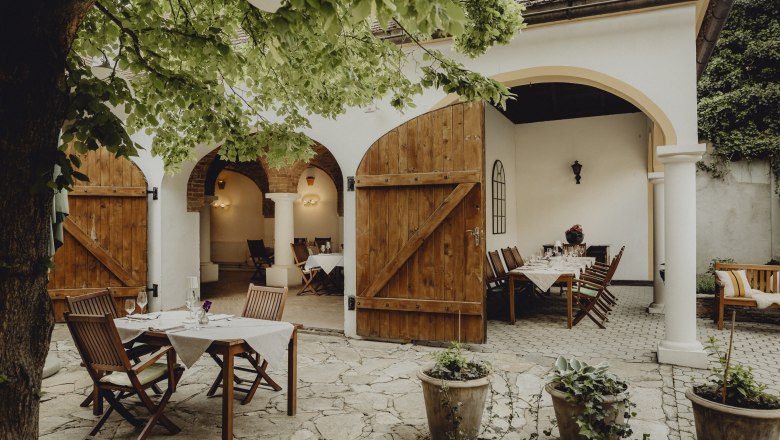 An inn to linger in, © Niederösterreich Werbung/Sophie Menegaldo Cozy guest garden with wooden tables and chairs under a tree, surrounded by white walls and open wooden doors.