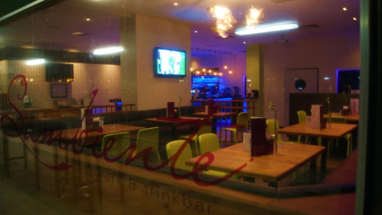 Local, © sambiente Interior view of an empty restaurant with wooden tables and colorful chairs, TV on the wall and illuminated bar in the background.