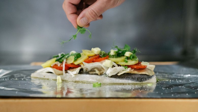 Culinary delight, © Felix Sommer A fish fillet with vegetables and herbs on aluminum foil, a hand adds garnish.