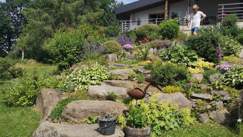 Watzl Albrechts show garden (06), © "Natur im Garten" A flowering rock garden with various plants and a metal sculpture of a bird. A person is standing on a terrace in the background.