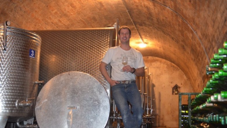 In the wine cellar, © Gass A man stands in a wine cellar next to large stainless steel tanks and bottles of wine.