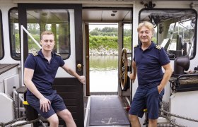 Event boat trip Haider, © Donau Niederösterreich, Barbara Elser Two men in naval uniform on a ship, with a view of a river and green shore landscape.