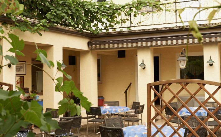 Guest garden at the Gasthof zum weissen Rössel, © Petra Schmidt A cozy guest garden with tables and chairs, surrounded by green leaves and a yellow building in the background.