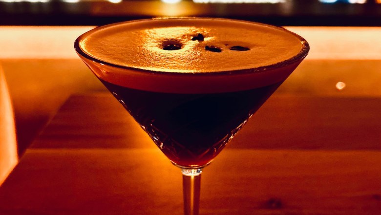 Bar drink, © Markus Jokesz A cocktail glass with a dark drink on a wooden table, a blurred bar in the background.