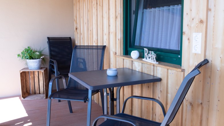 Balcony for a cozy stay, © Familie Schwalm A balcony with wooden walls, two black chairs, a table and a plant in a pot.