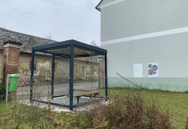 Bus stop at the parish garden, © Marktgemeinde Königsbrunn A modern bus stop with glass walls next to an old building and a green meadow.