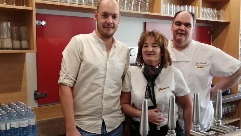 Host family father, mother, son behind the bar, © Gasthubers Wirtshaus Three people stand behind a bar with taps and glasses in the background.
