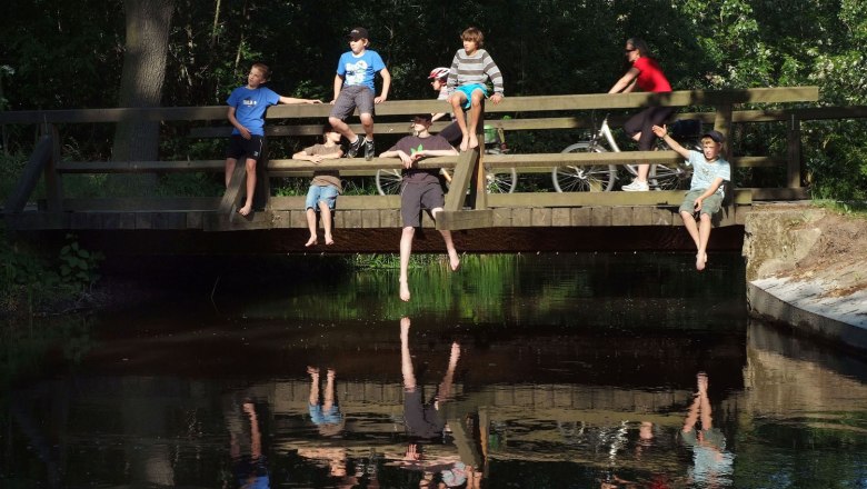 Lindenhof, © Seymannfilm People sit on a wooden bridge over a river, some with their legs dangling. One person is riding a bicycle across the bridge.