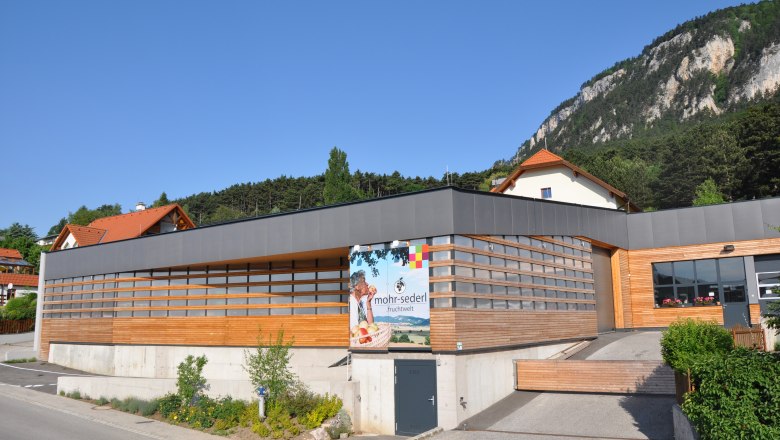 Mohr-Sederl Fruit World, © Mohr-Sederl Fruchtwelt Modern building with wooden cladding and advertising sign for Mohr-Sederl Fruchtwelt, surrounded by green countryside and mountains.