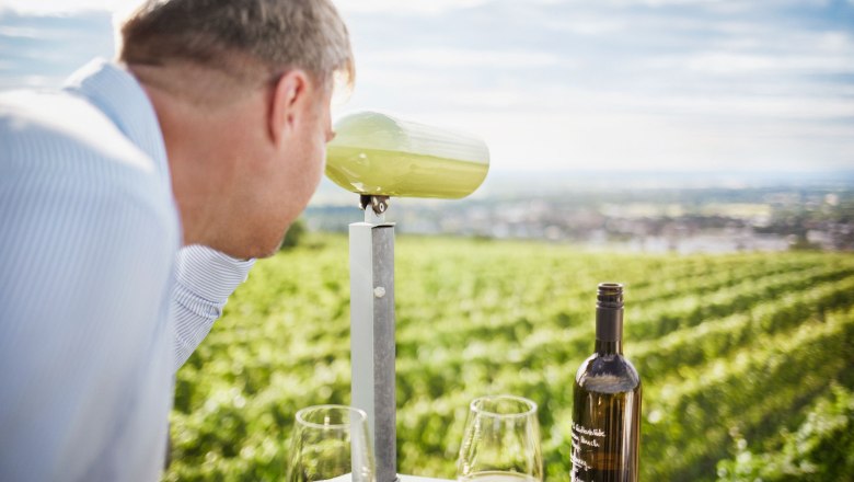 Wine trail Langenlois, © © Waldviertel Tourismus, Gerhard Wasserbauer A man looks through binoculars at a field of vines. There is a bottle of wine and two glasses on the table.