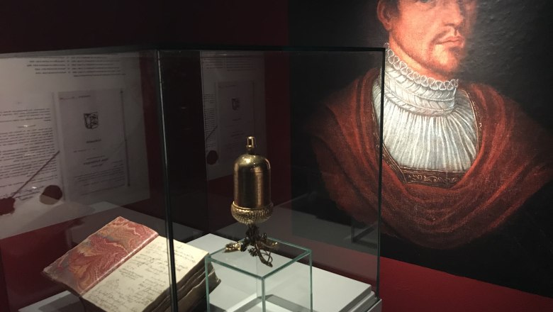 The Corvinus cup in the PIZ1000, © Marktgemeinde Pitten Exhibition with a golden cup, an old book and a portrait in a museum.