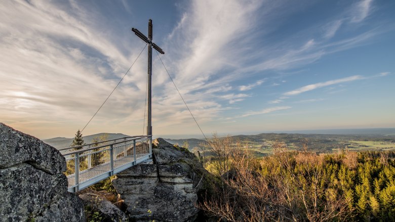 Excursion paradise Nebelstein, © Christian Freitag A summit cross on a rock with a viewing platform, surrounded by forest and a wide view over the landscape under a blue sky.
